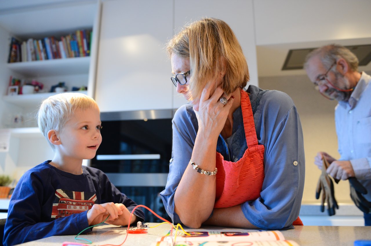 A young boy and a woman work on a simple electrical circuit together, while an older man stands nearby.