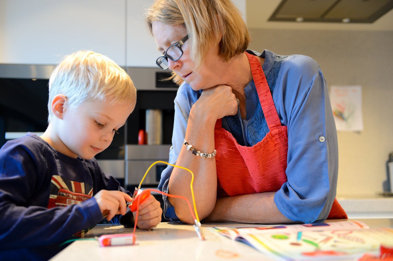 A young boy connects wires to a battery while an older woman watches closely in a kitchen setting.