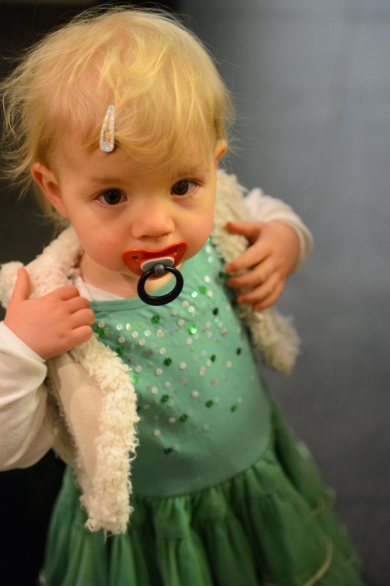 A young child with a pacifier looks at the camera, wearing a green dress and a fluffy white vest.