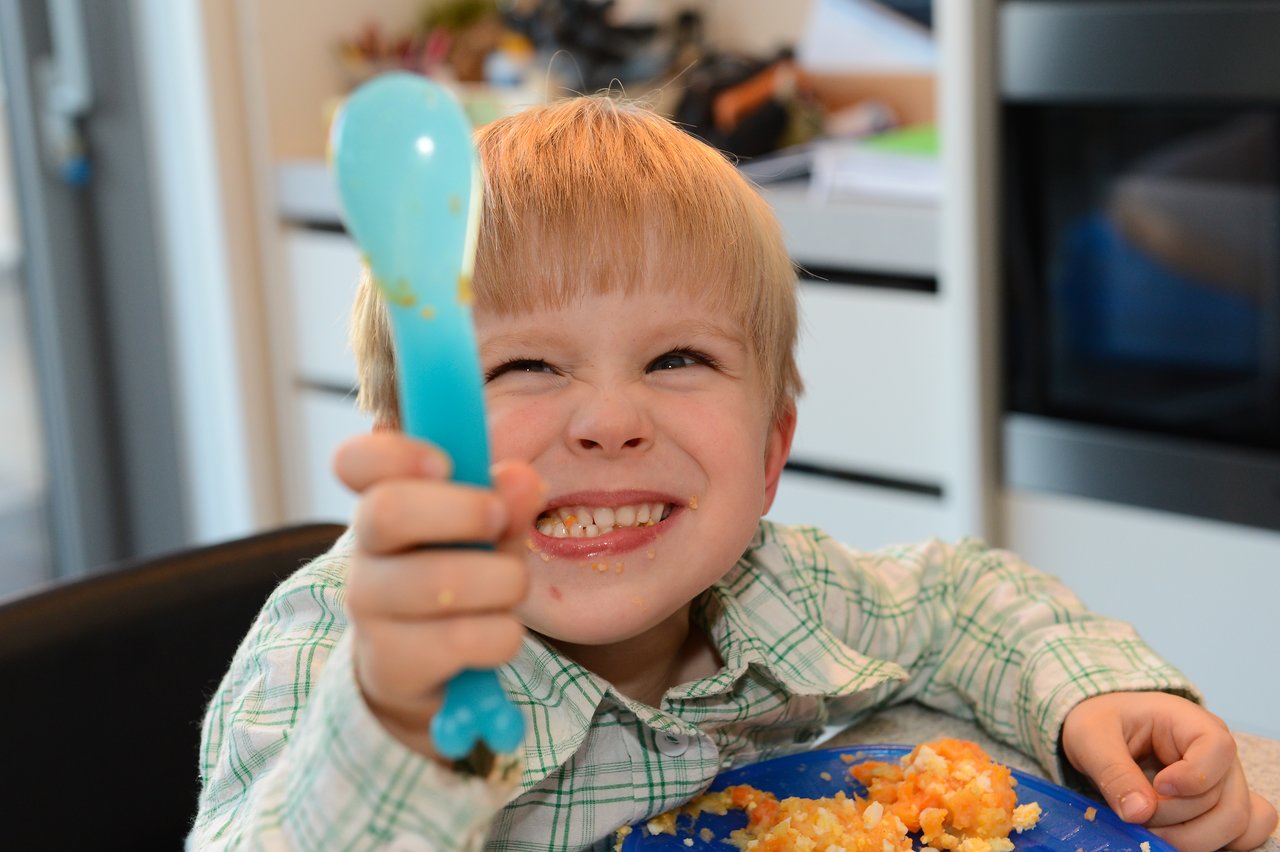 A young child in a plaid shirt smiles and holds up a blue spoon while eating a meal.