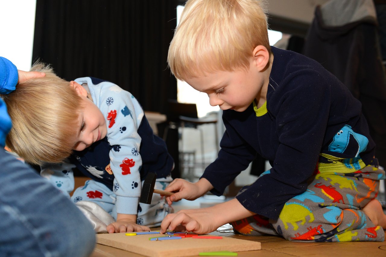 Two young children in pajamas play on the floor, one hammering colorful pieces onto a wooden board.