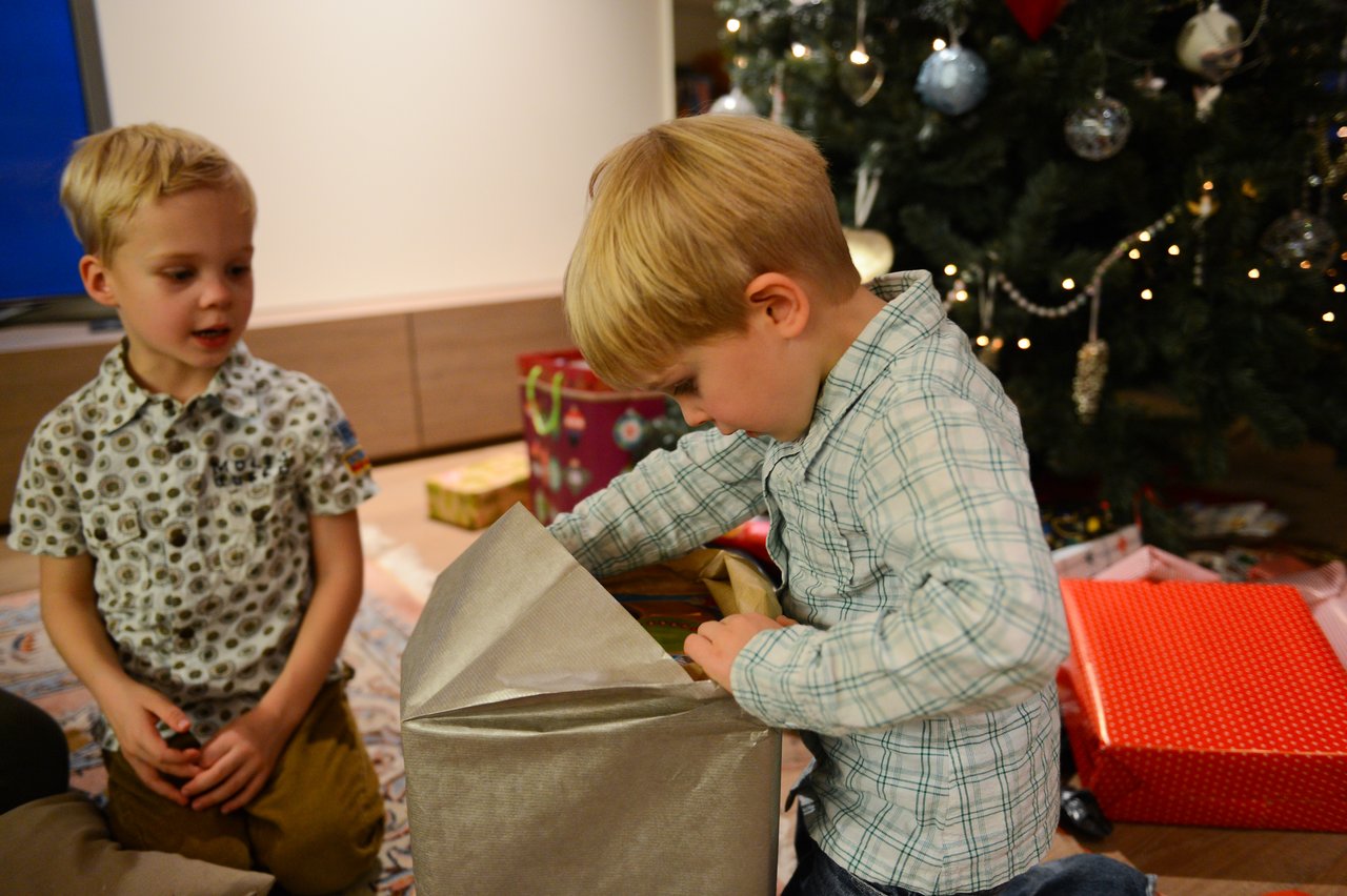 A child in a plaid shirt eagerly unwraps a gift while another child watches near a decorated Christmas tree.