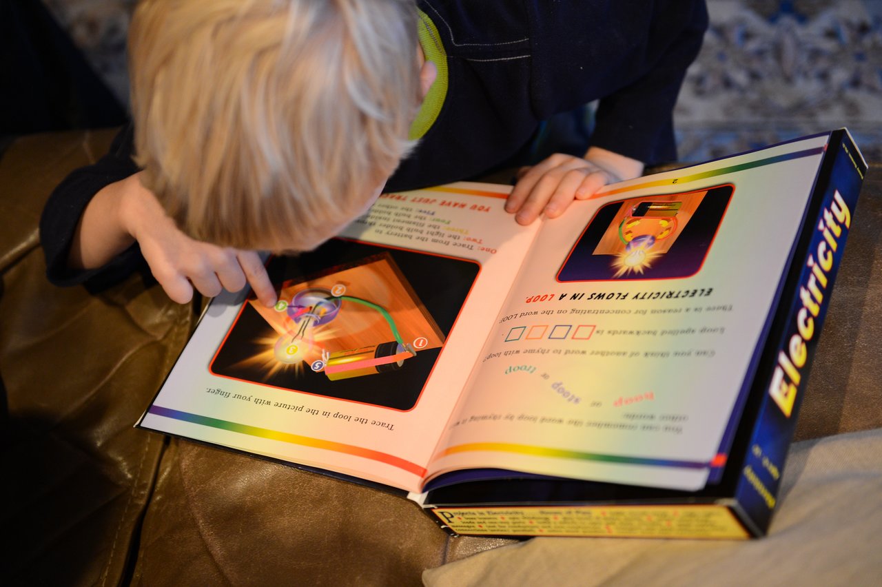 A child closely examines a book about electricity, pointing at an illustration of a circuit.
