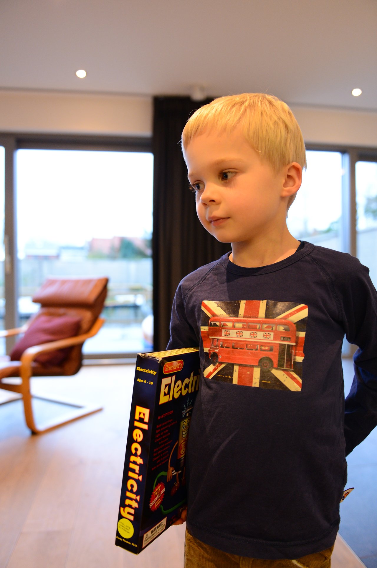 A young child holds a newly unwrapped electricity-themed toy while standing in a living room.