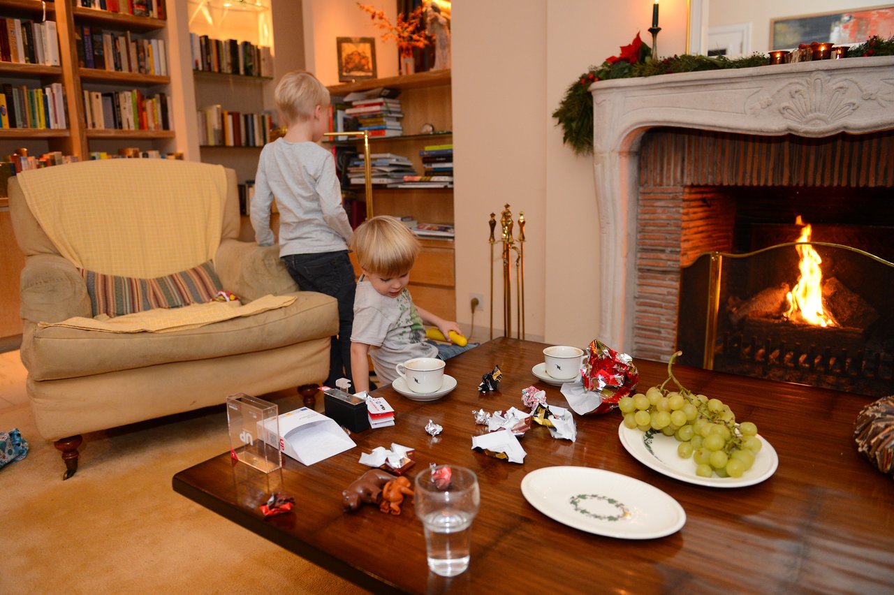 Two young children stand near a table with snacks and drinks in a cozy living room with a fireplace.