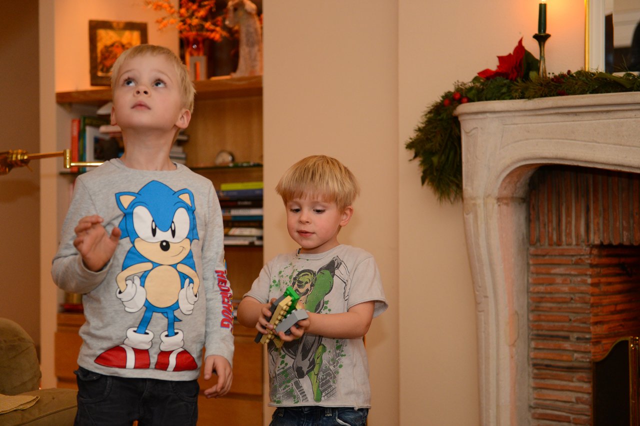 Two young boys stand near a decorated fireplace, one holding a toy while the other looks up.
