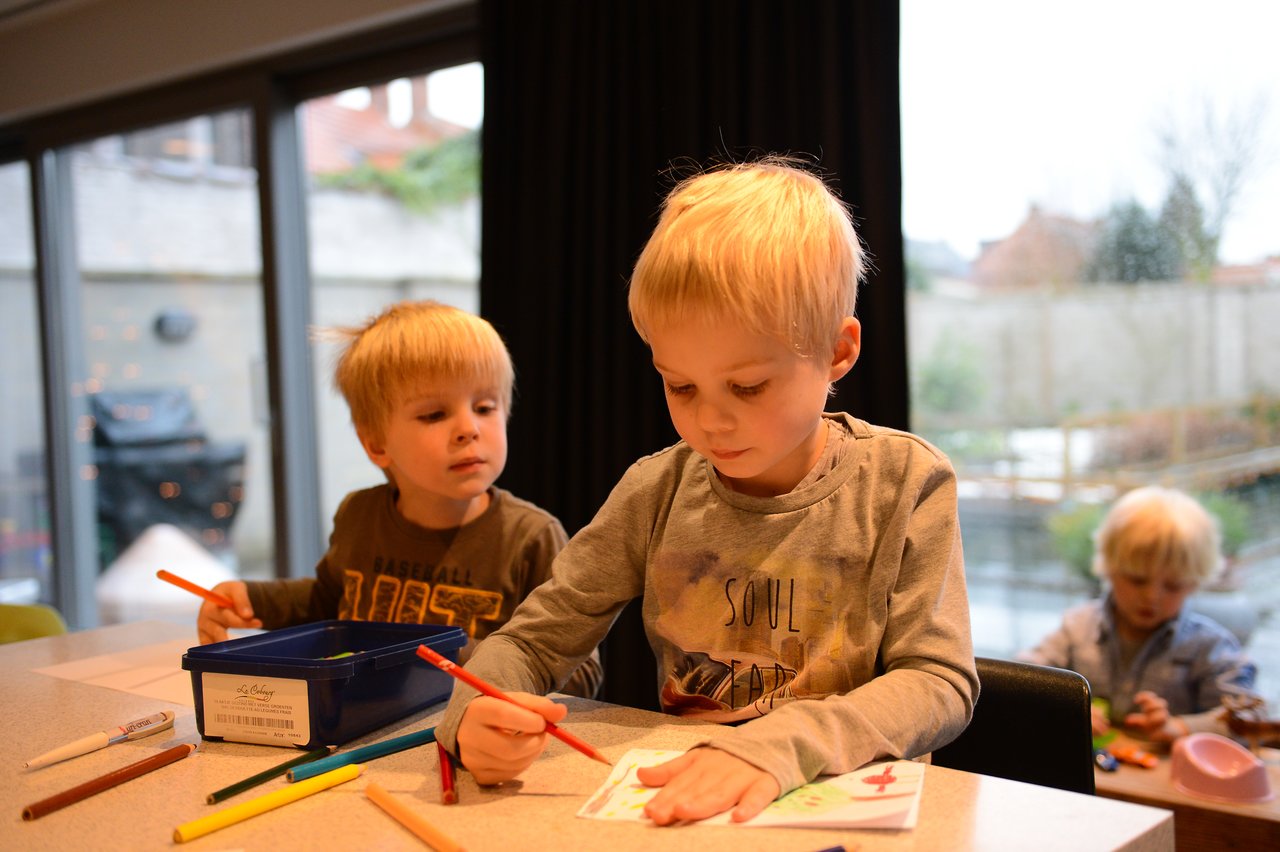 Two young children sit at a table, coloring with markers and pencils, while another child plays in the background.