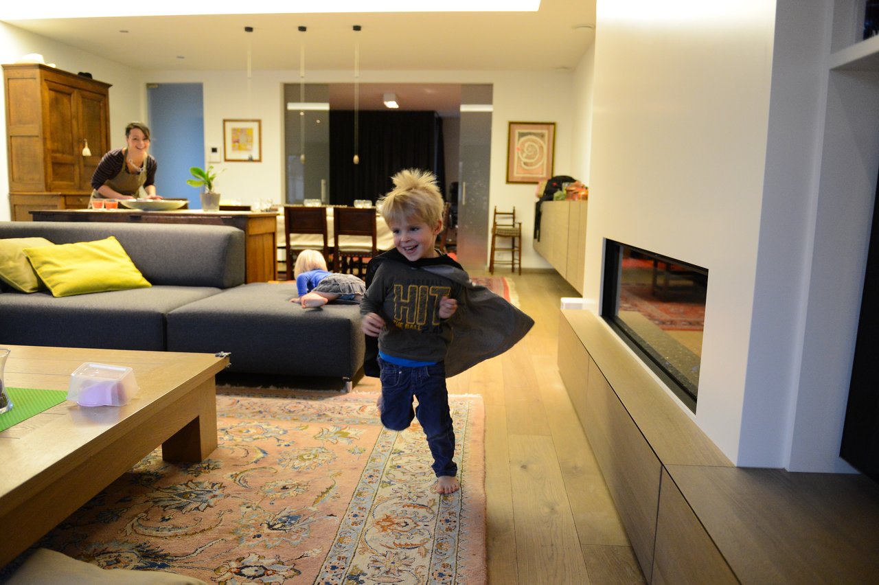 A young child runs barefoot through the living room, wearing a cape, while others engage in activities nearby.