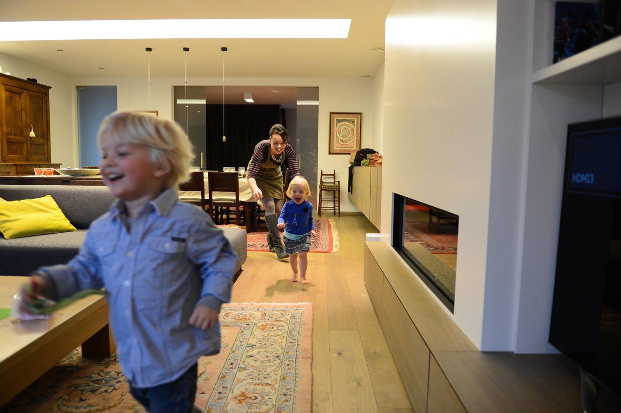 Two young children run through a living room, laughing, while a woman playfully chases them from behind.