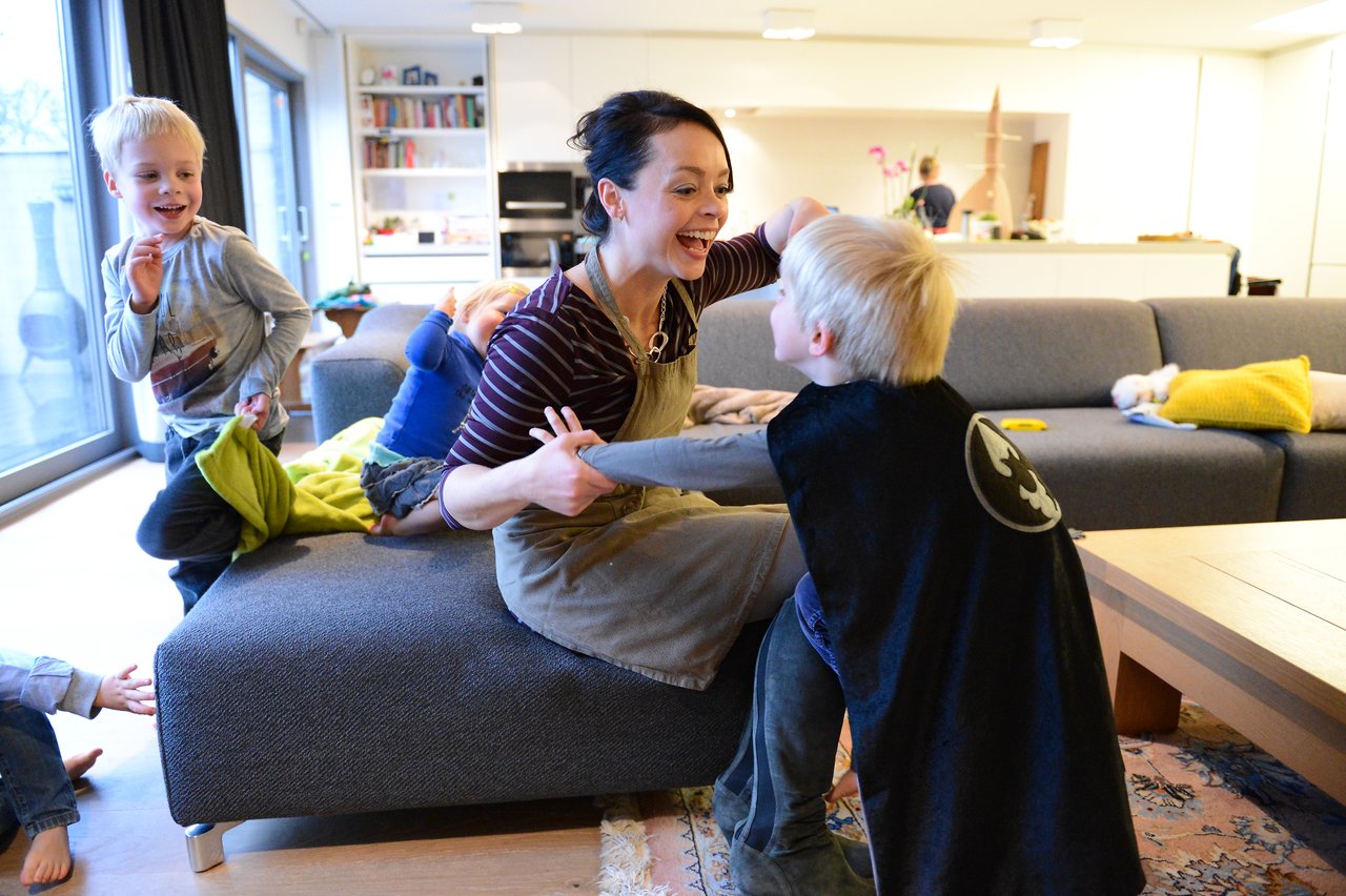 A woman and a child in a Batman cape playfully interact while other children watch and laugh in a living room.