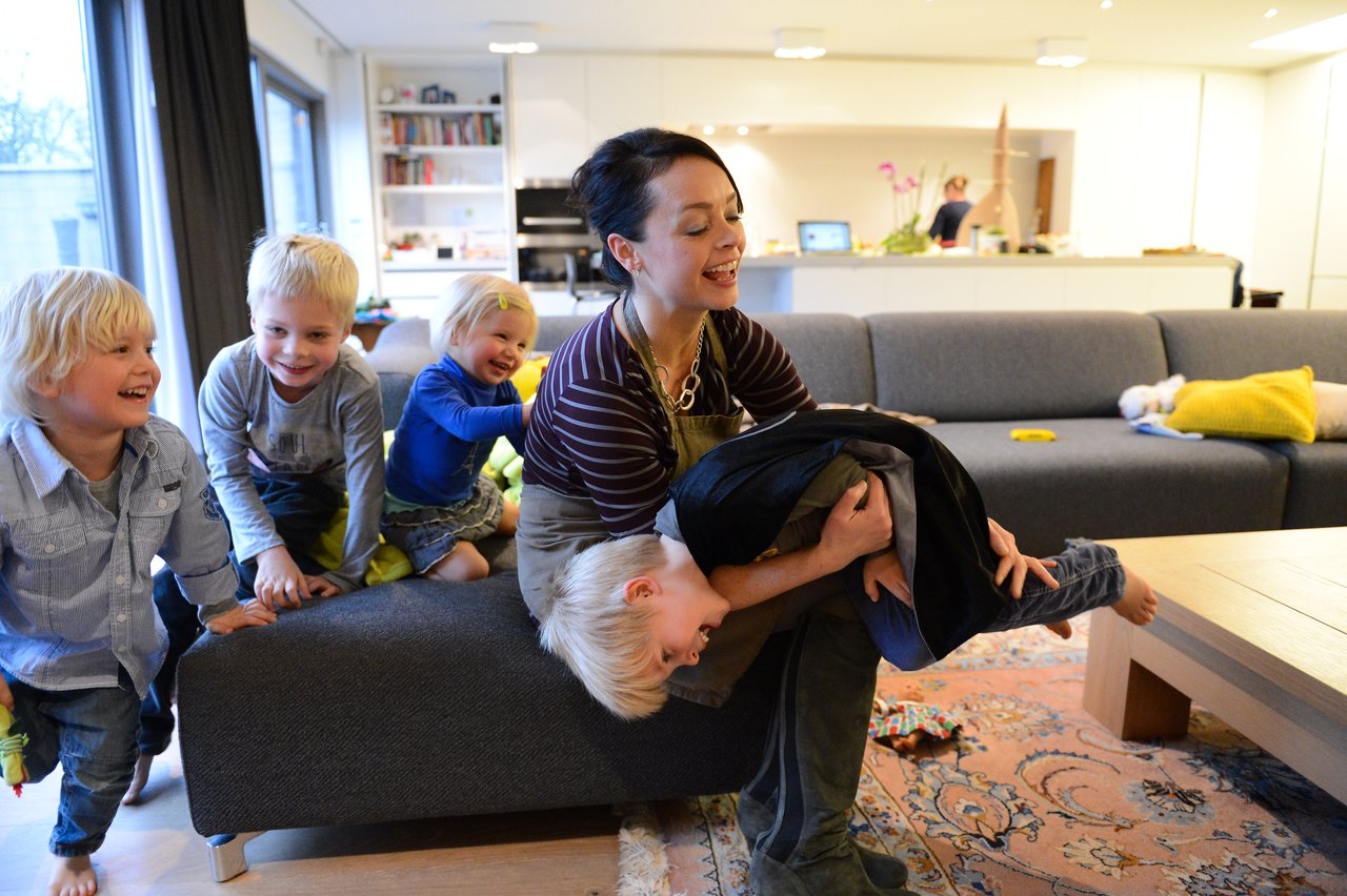 A woman playfully holds a child upside down while other children laugh and watch from the couch.