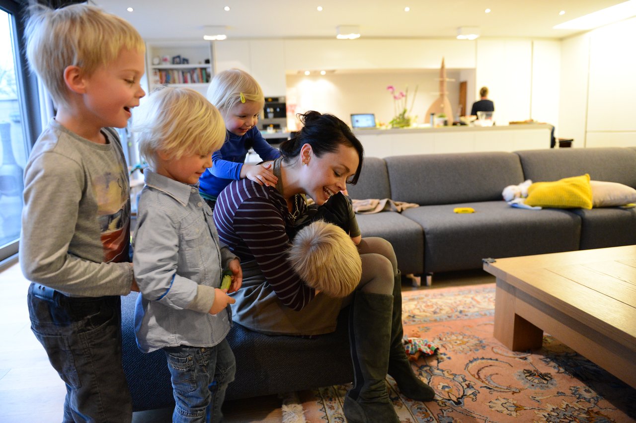 A woman sits and smiles while hugging a child, surrounded by three other children in a cozy living room.