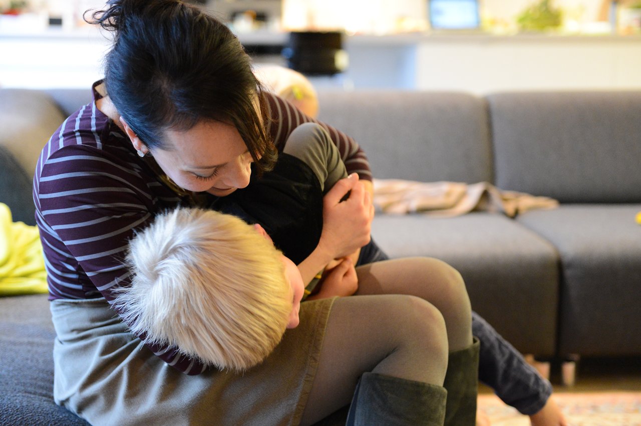 A woman sits on a couch, hugging and playfully holding a child with blonde hair in her arms.