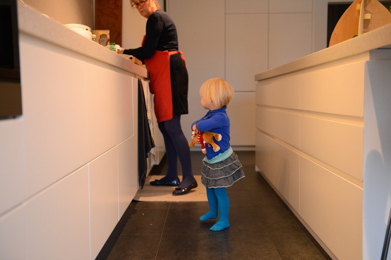 A child holding a stuffed toy stands in the kitchen while an adult prepares food at the counter.
