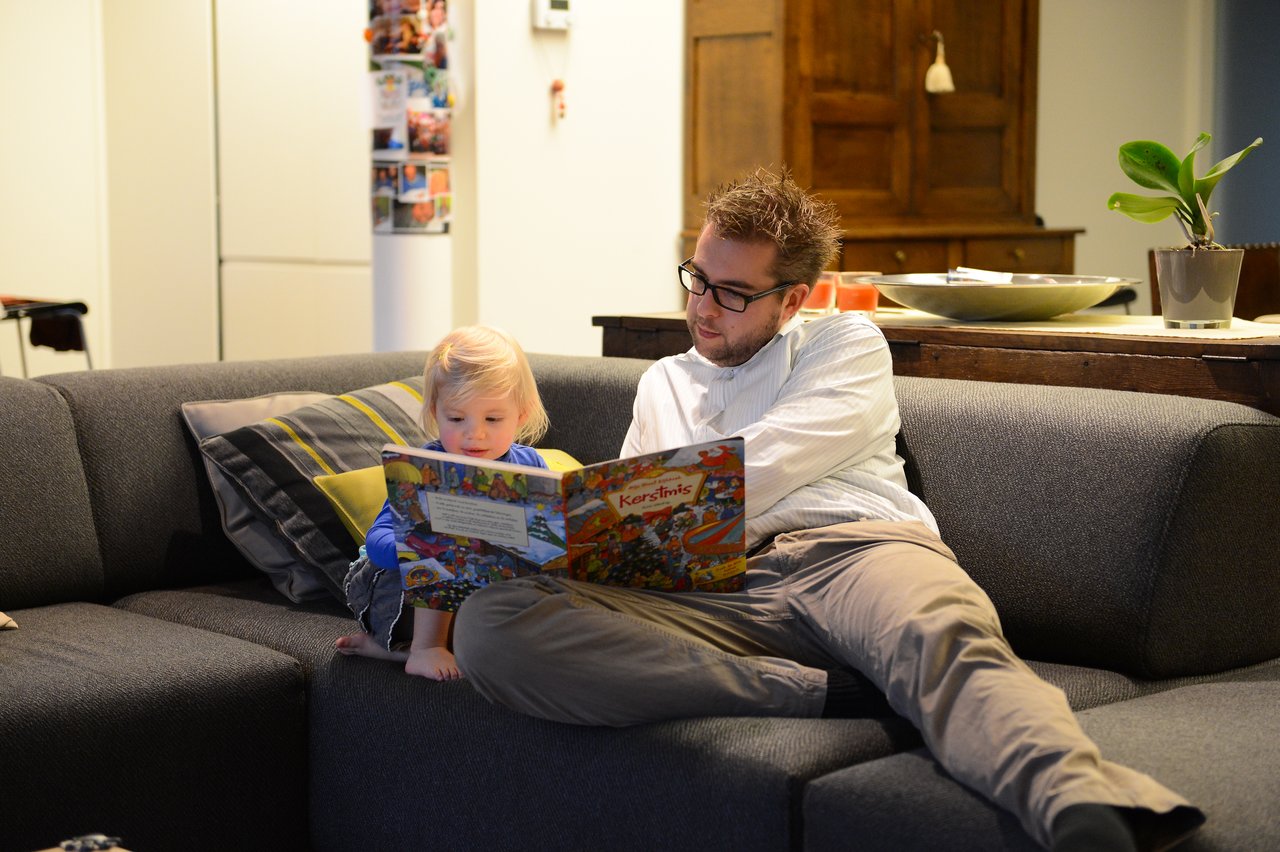 A man and a young child sit on a couch, reading a Christmas-themed book together.
