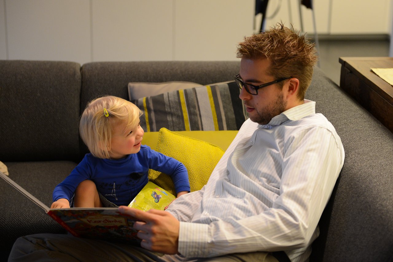 A man and a young child sit on a couch, reading a book together and smiling.