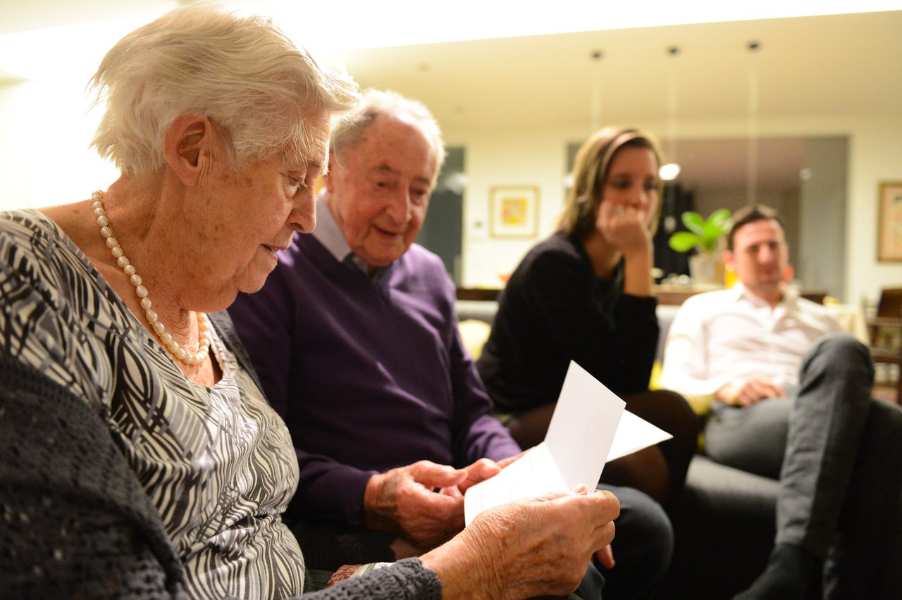 An elderly woman reads a card while sitting with family members in a warmly lit living room on Christmas Eve.