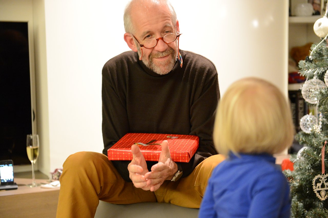 An older man holding a wrapped gift smiles while talking to a young child near a decorated Christmas tree.