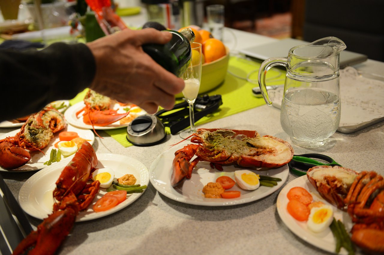 A person pours champagne into a glass beside plates of lobster, eggs, and vegetables on a festive table.
