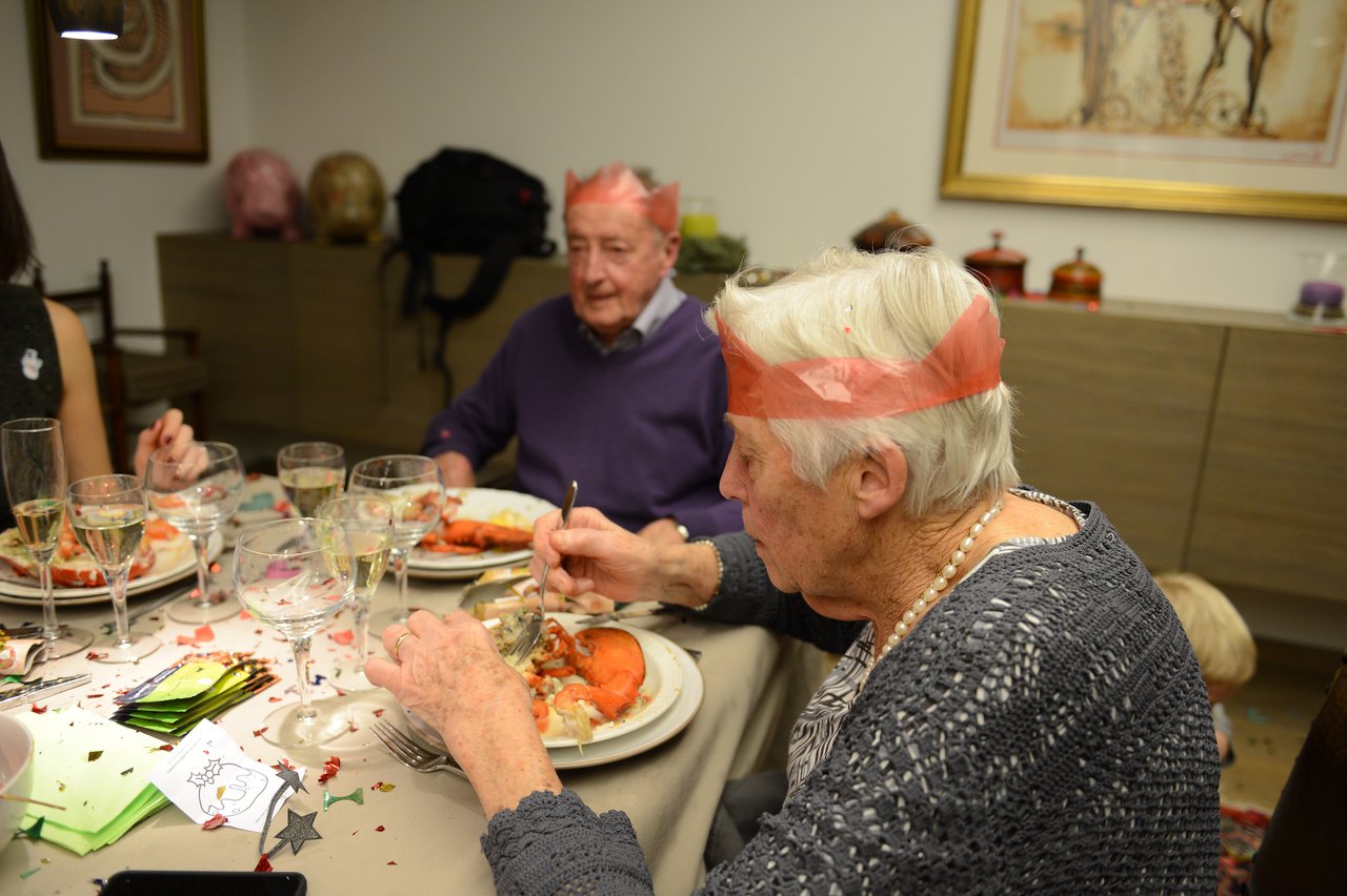 Elderly woman and man wearing paper crowns enjoy a festive Christmas Eve dinner with lobster and champagne.