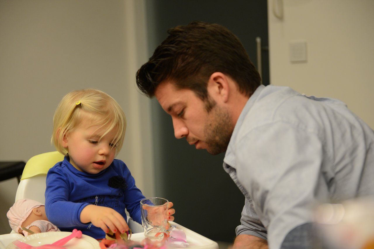 A young child in a high chair plays with toys while an adult leans in to engage with them.