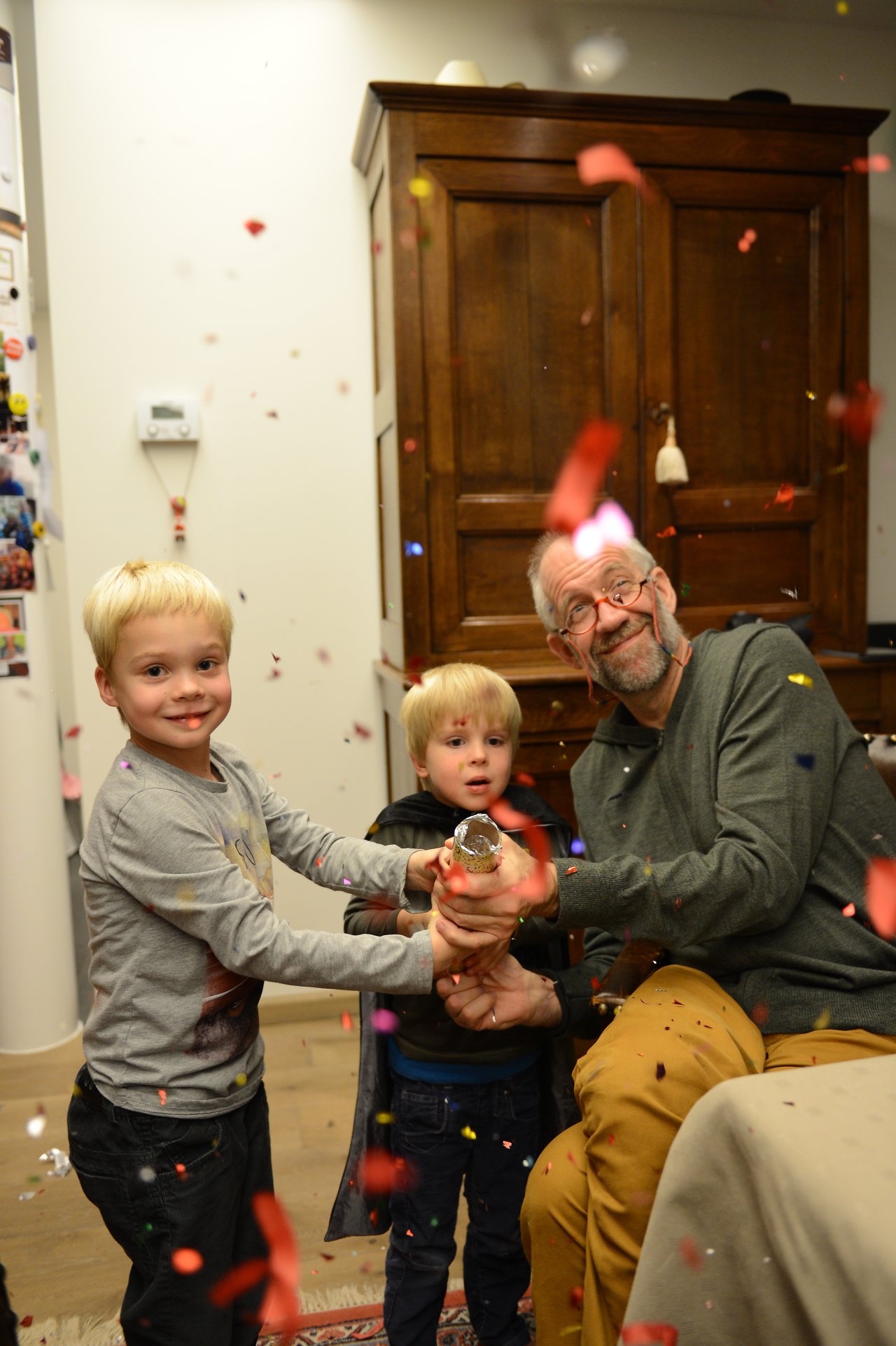 Two children and an older man pop a party cracker, releasing colorful confetti in a festive indoor setting.