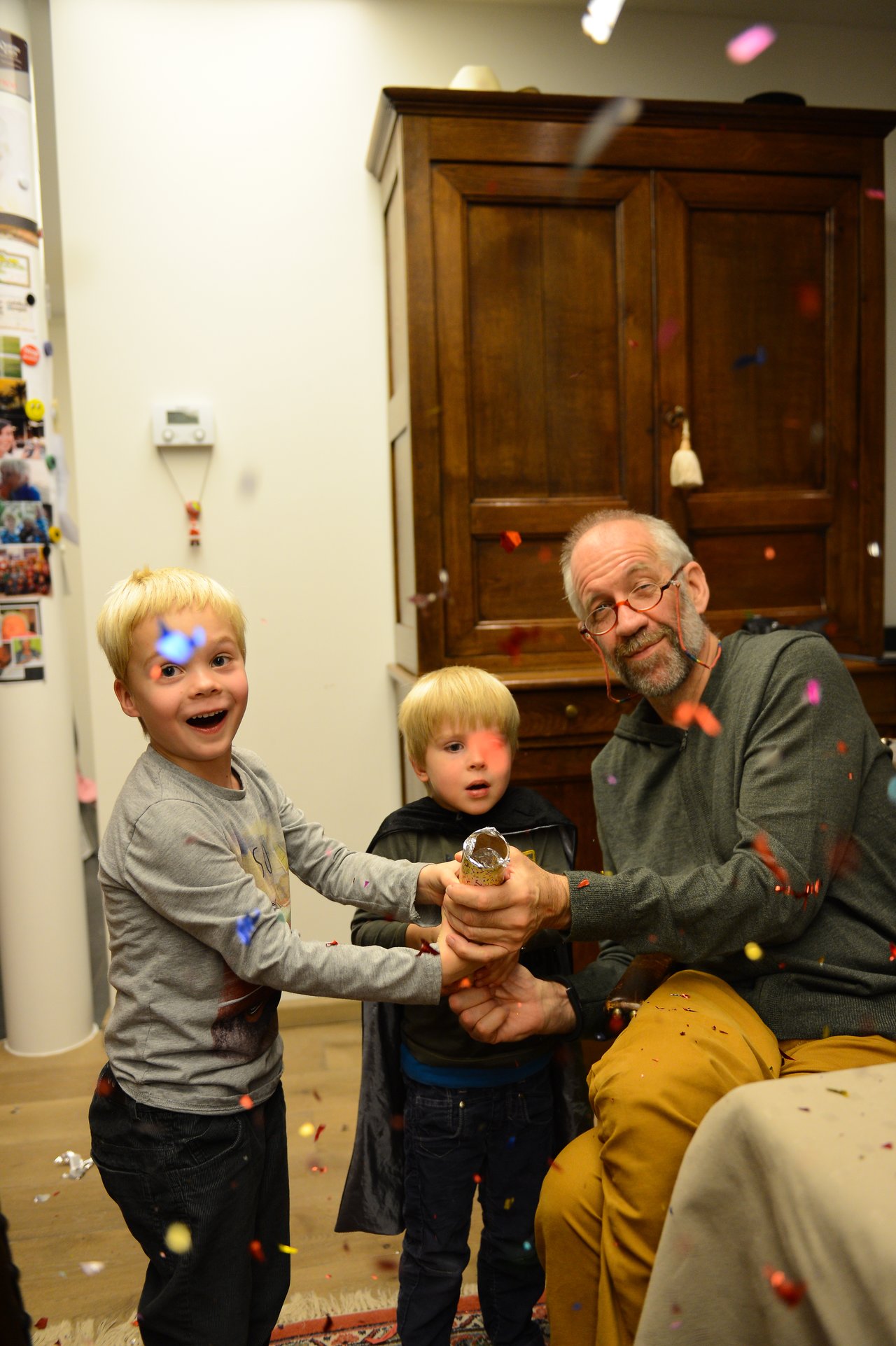 A man and two children pop a party cracker, releasing colorful confetti in a festive indoor setting.