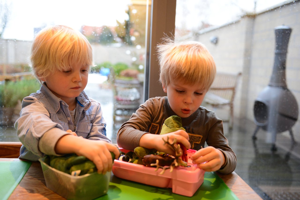 Two young children sit at a table, focused on playing with vegetables and small objects in plastic containers.