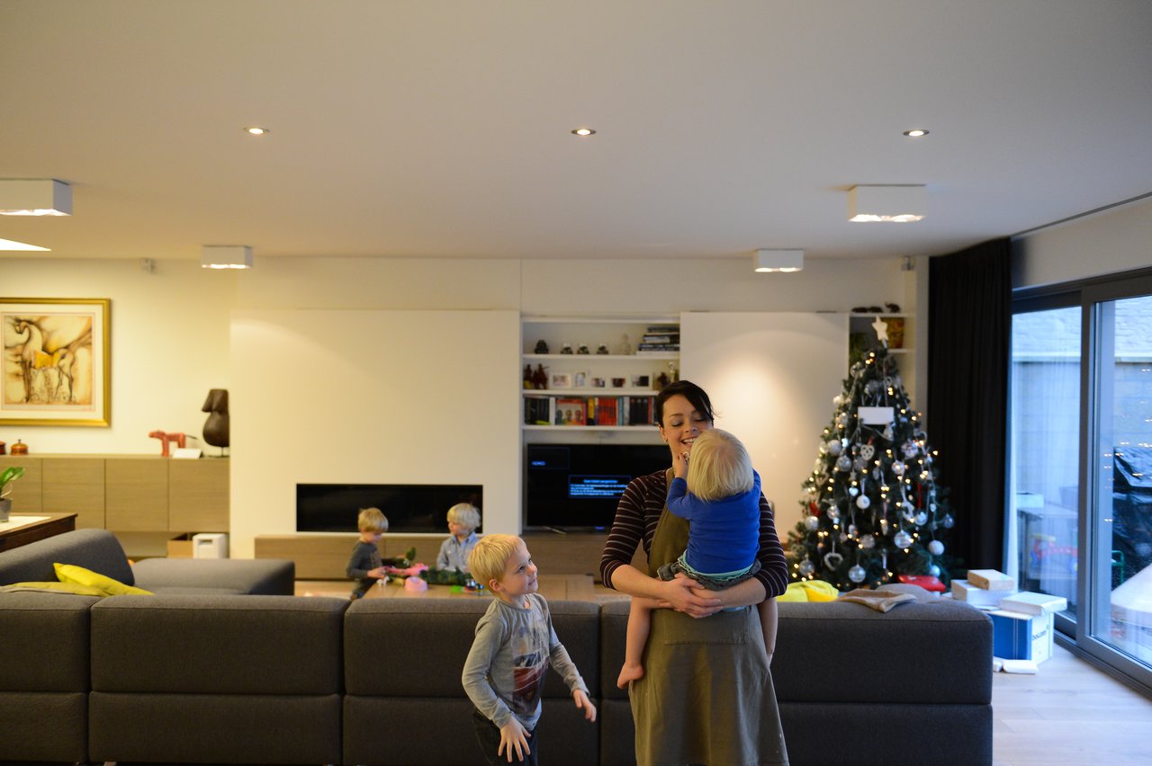 A woman holds a young child while another child stands nearby in a living room with a Christmas tree.