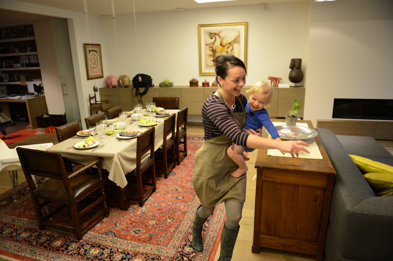 A woman in an apron holds a smiling child while setting a table for Christmas Eve dinner.