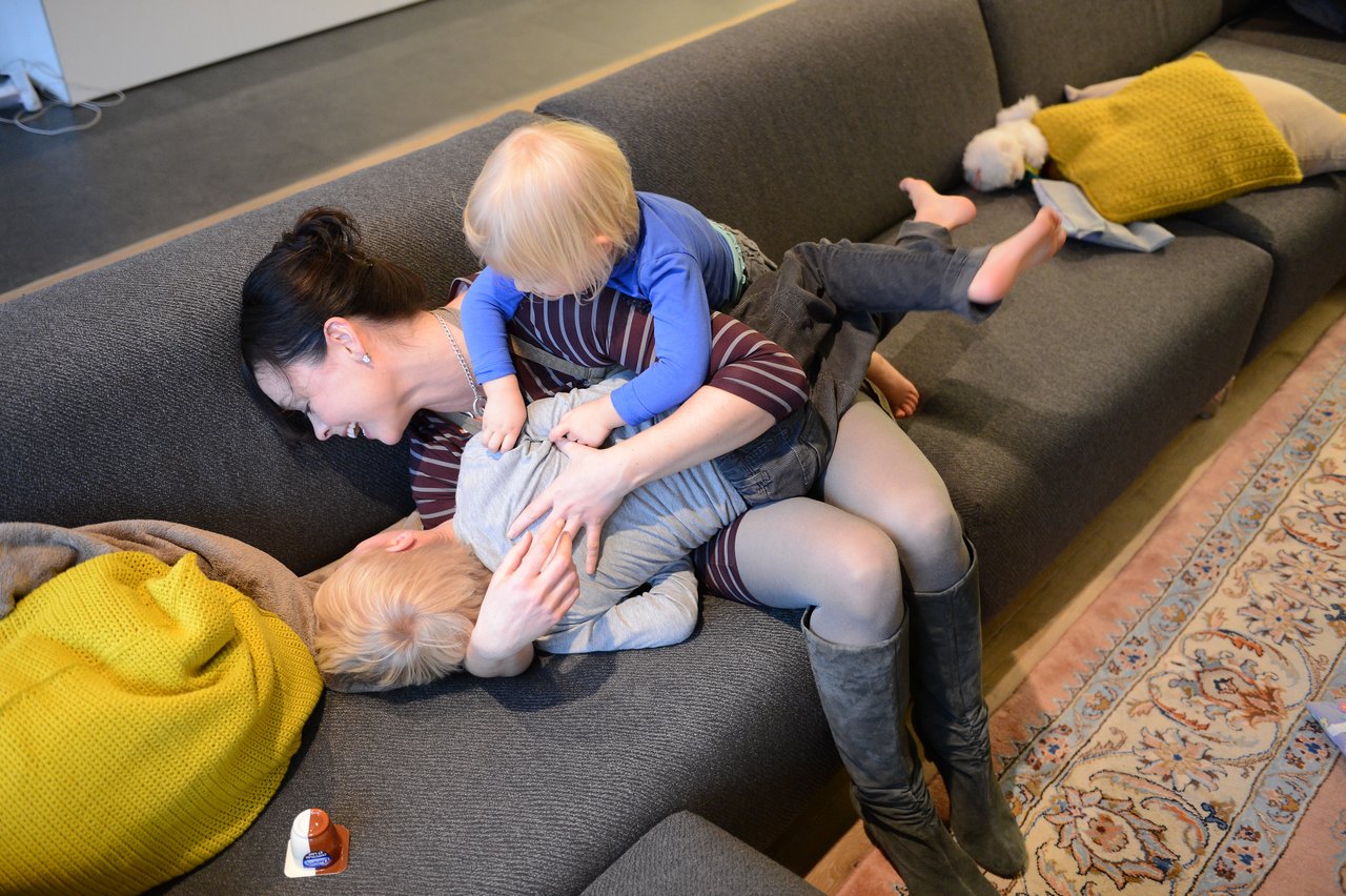 A woman playfully wrestles with two young children on a gray couch, all laughing and enjoying the moment.