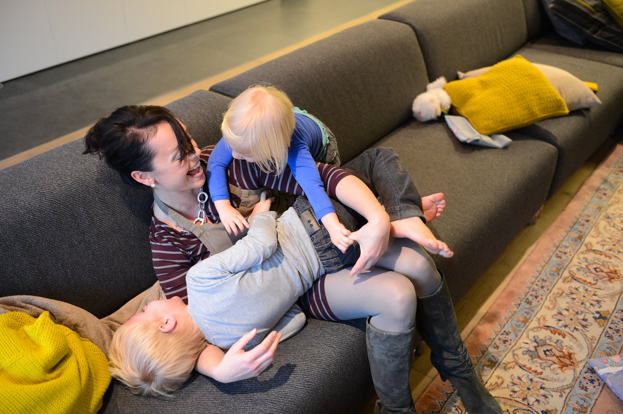 A woman sits on a couch, smiling while playfully holding two young children climbing on her.