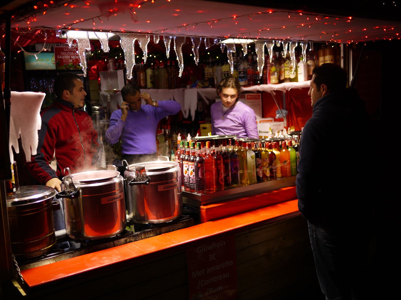 A customer orders a drink at a Christmas market stall in Antwerp, with steaming pots and bottled beverages displayed.