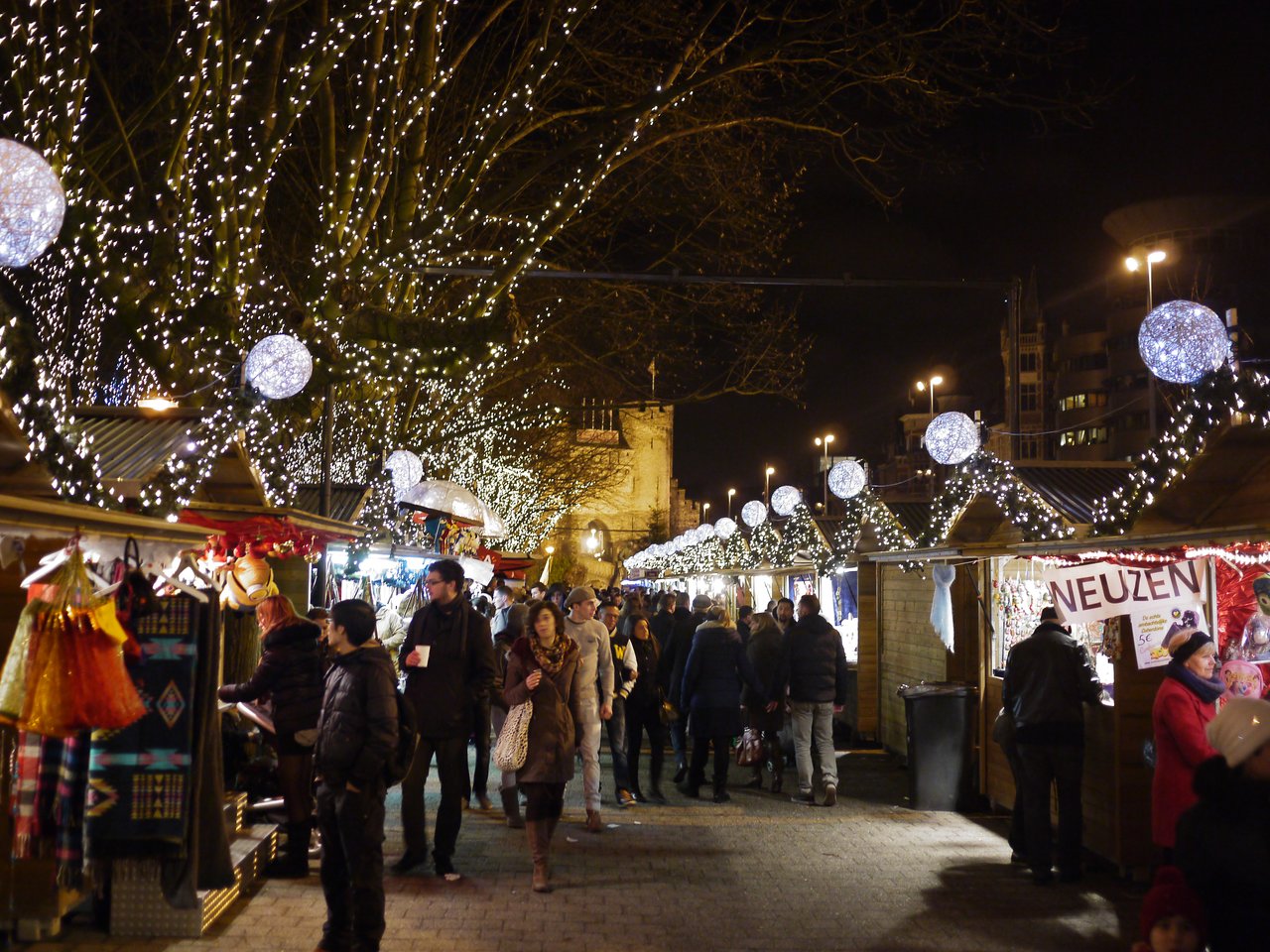 People walk through a festive Christmas market in Antwerp, browsing wooden stalls decorated with lights and ornaments at night.