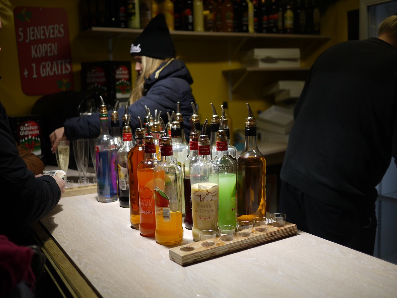 A market stall in Antwerp selling various flavored spirits, with people ordering drinks and a bartender preparing them.