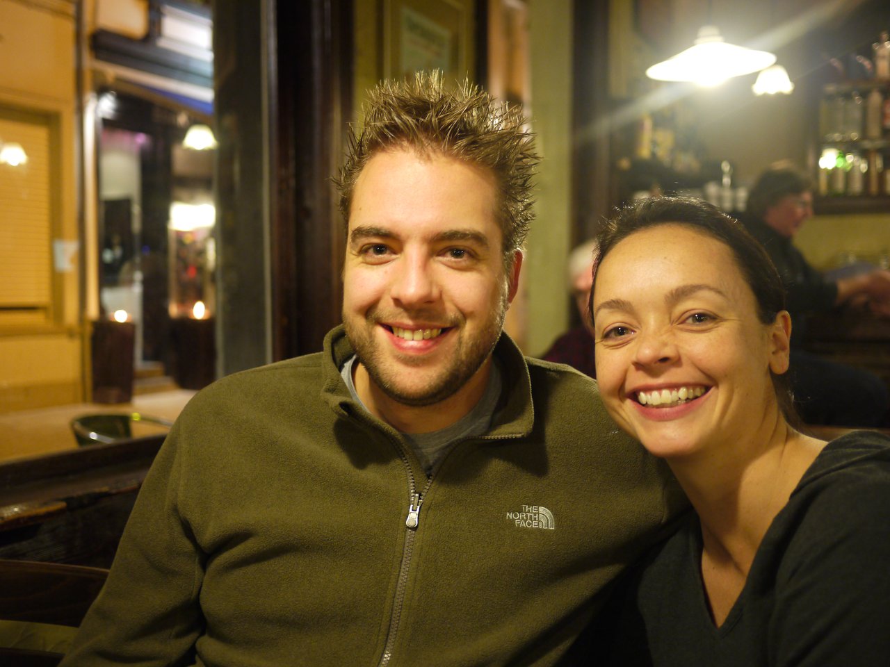 A smiling man and woman sitting together in a warmly lit indoor setting at the Christmas market in Antwerp.