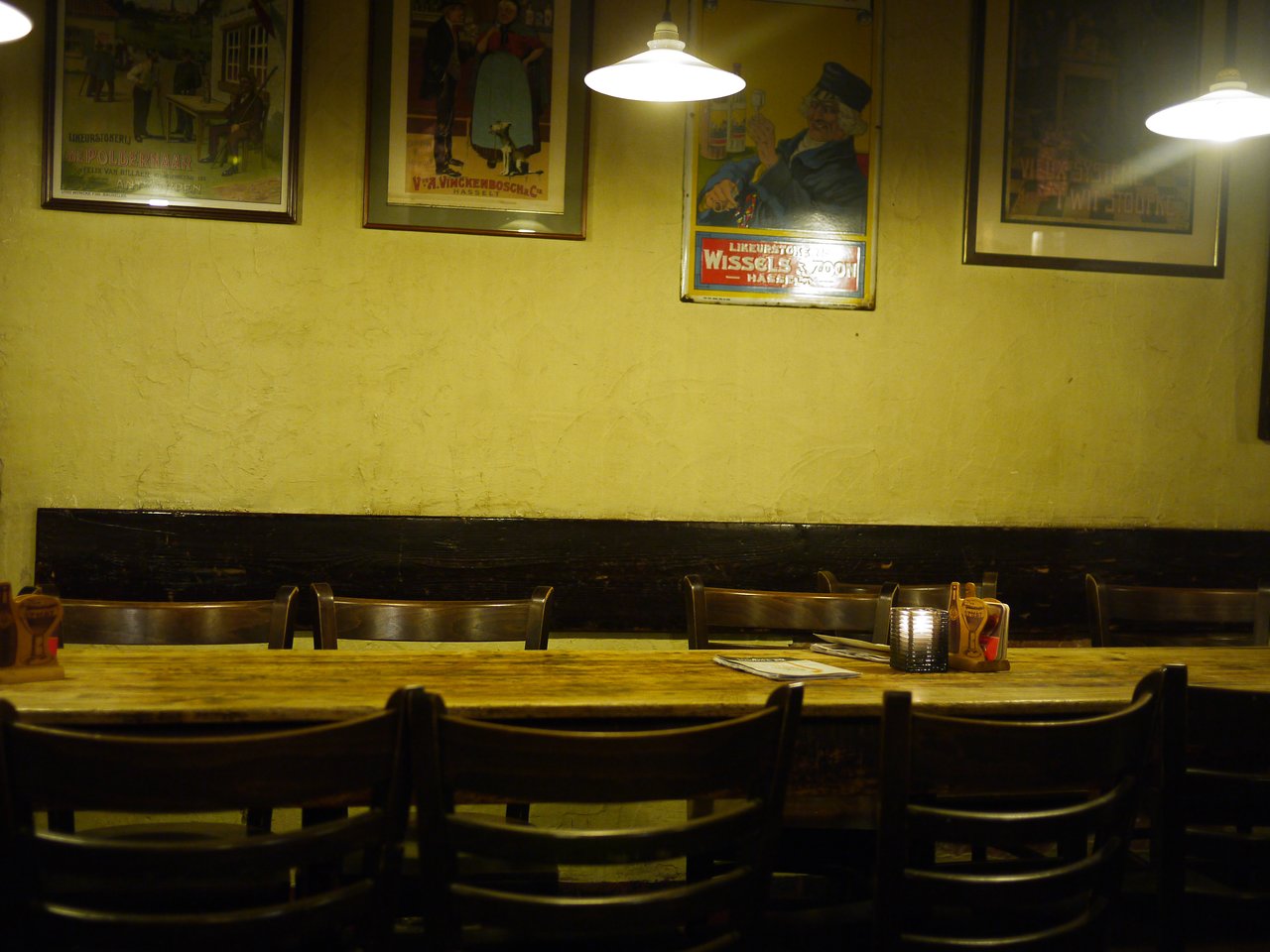 A long wooden table with chairs in a dimly lit restaurant, decorated with vintage posters and a candle.
