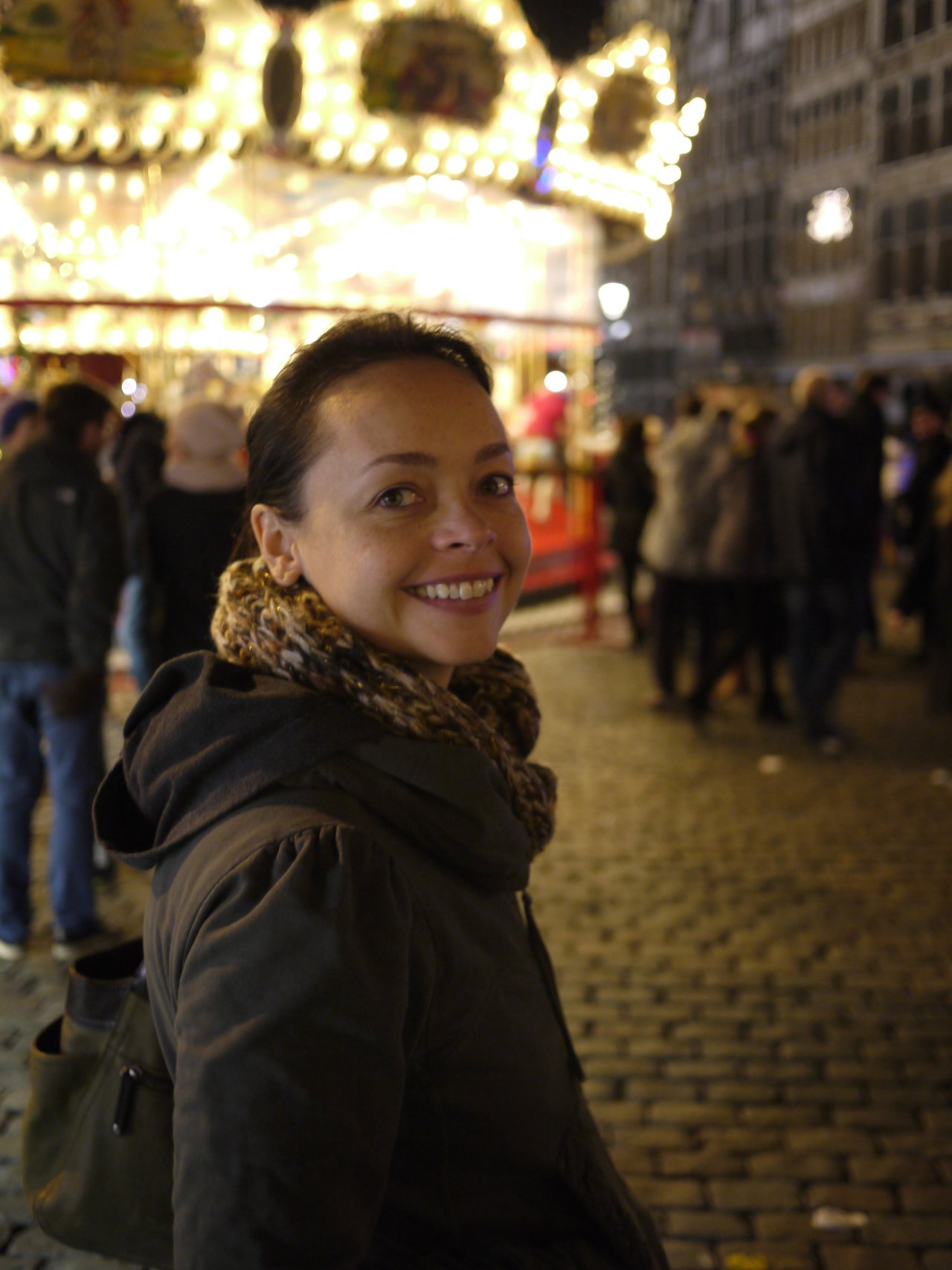 A woman in a winter coat smiles at the camera in a busy Christmas market with bright lights.