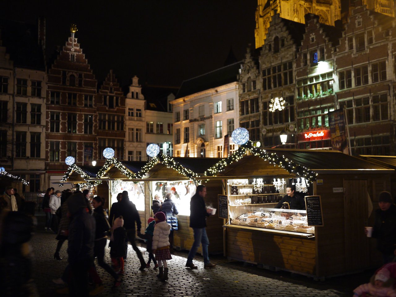 People walk through a Christmas market in Antwerp at night, with wooden stalls selling food and holiday items.