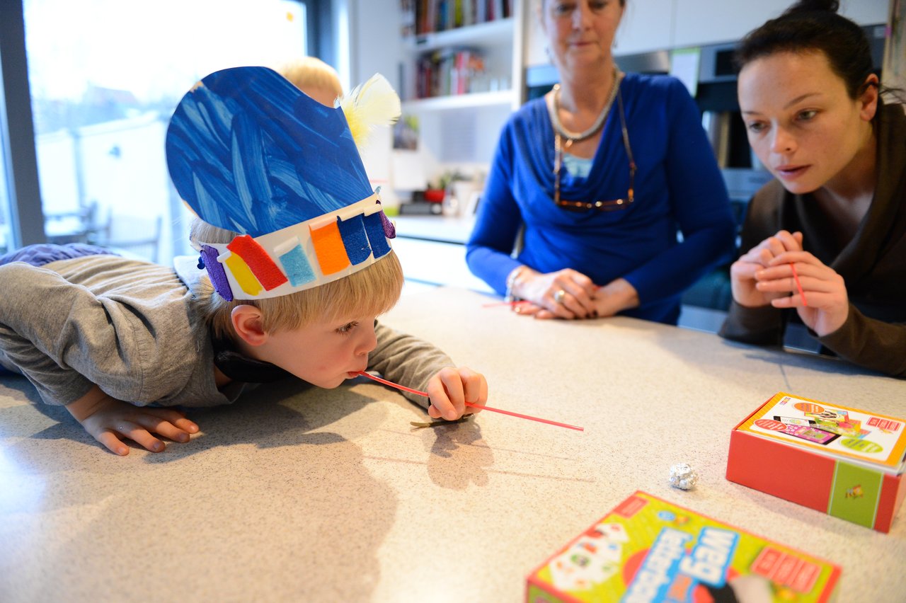 A child wearing a colorful paper hat blows through a straw, moving a small object, while others watch closely.