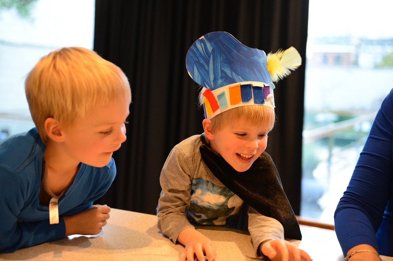 Two young children play at a table, one wearing a colorful handmade hat and smiling excitedly.