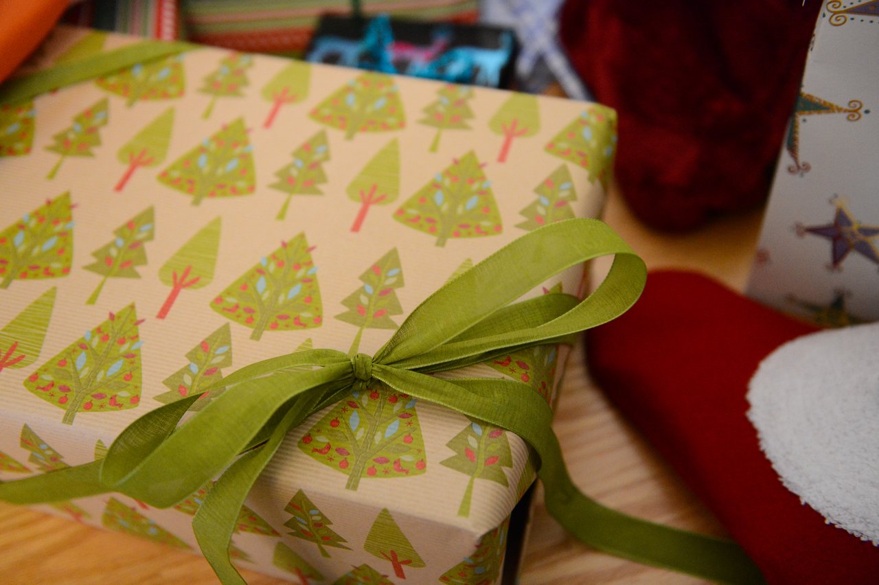 A wrapped Christmas gift with tree-patterned paper and a green ribbon, placed among other presents on a wooden surface.
