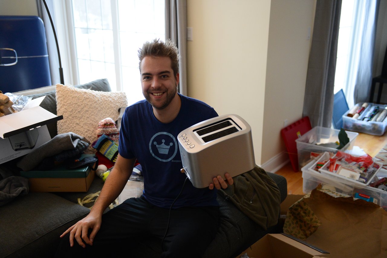 A smiling person sits on a couch, holding a new toaster among opened gifts and wrapping materials.