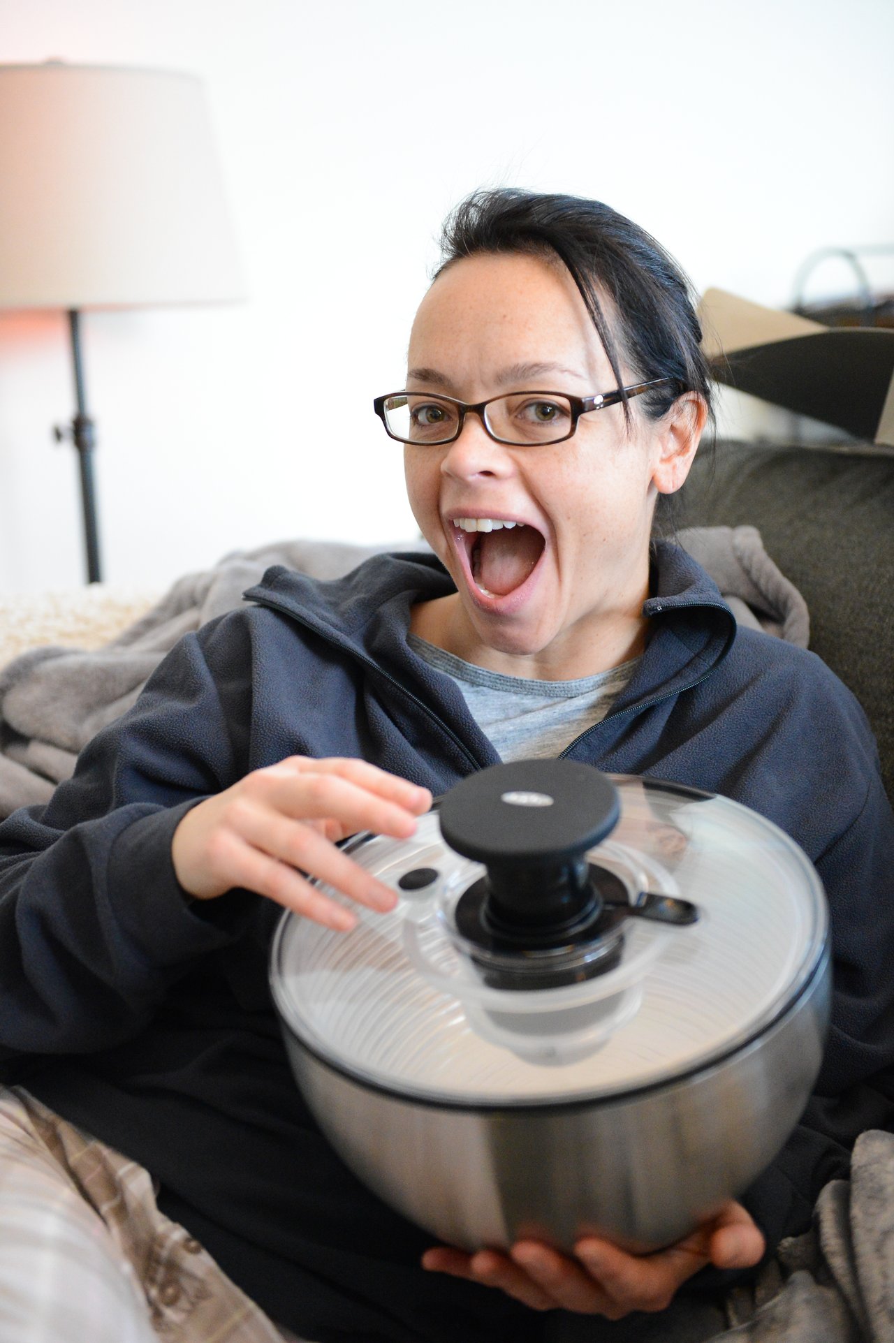 A person with glasses smiles excitedly while holding a new salad spinner, sitting on a couch.