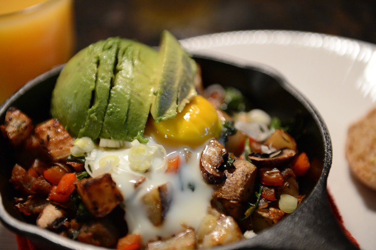 A skillet with roasted vegetables, a poached egg, and sliced avocado, served with a side of bread.