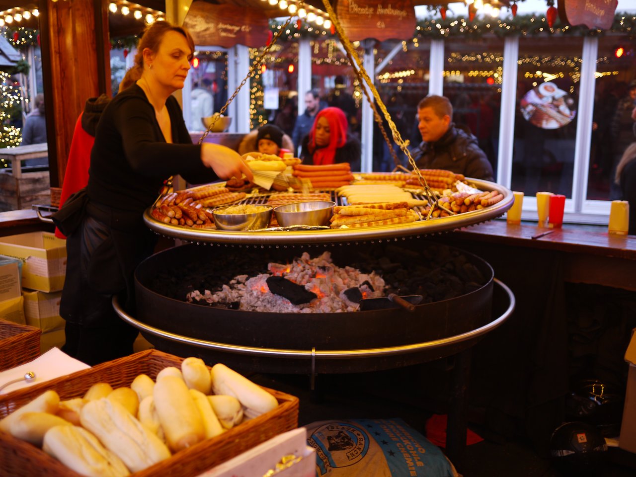 A woman grills sausages and bread over an open fire at a festive market stall, serving customers.