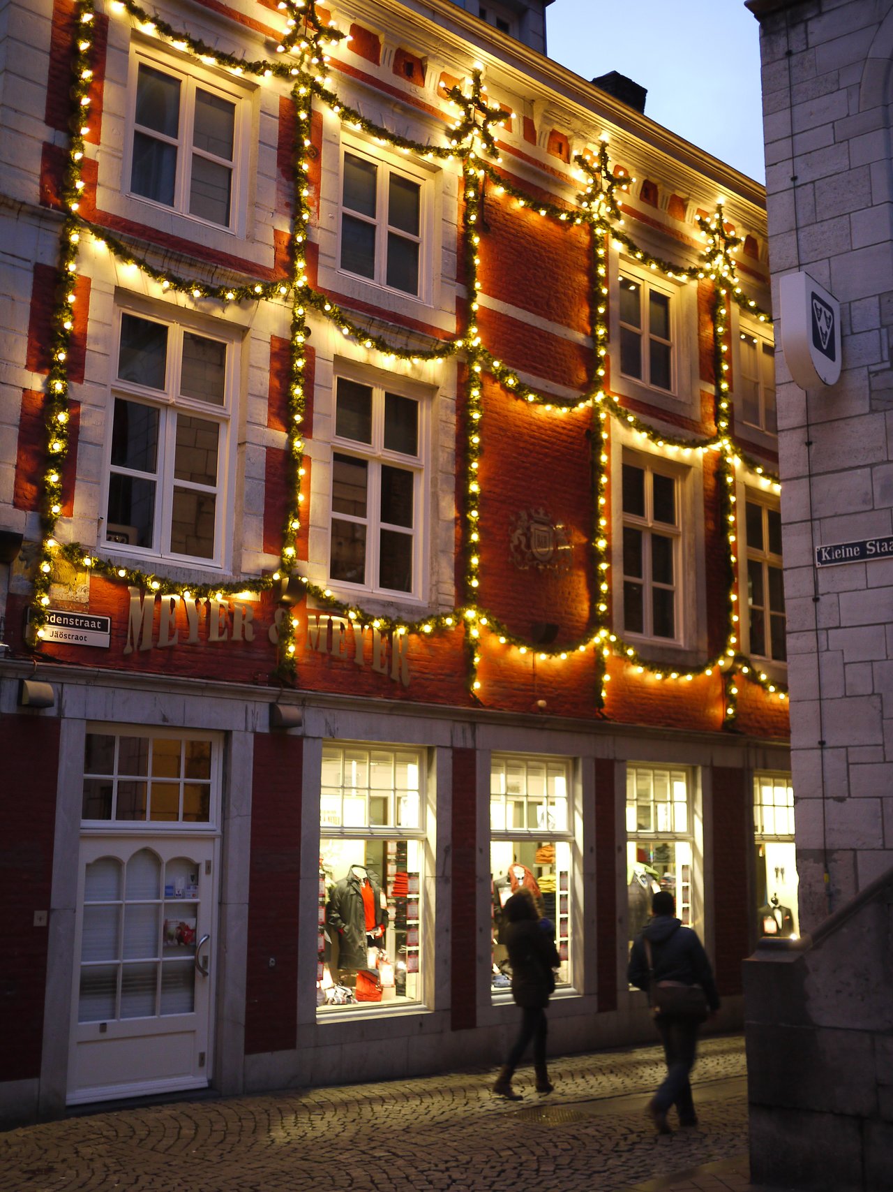 A brick building decorated with Christmas lights, with two people walking past illuminated shop windows in the evening.