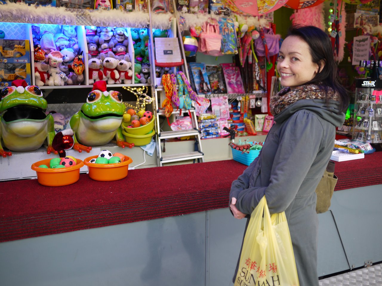 A woman in a coat smiles while holding a shopping bag at a colorful market stall with toys and decorations.