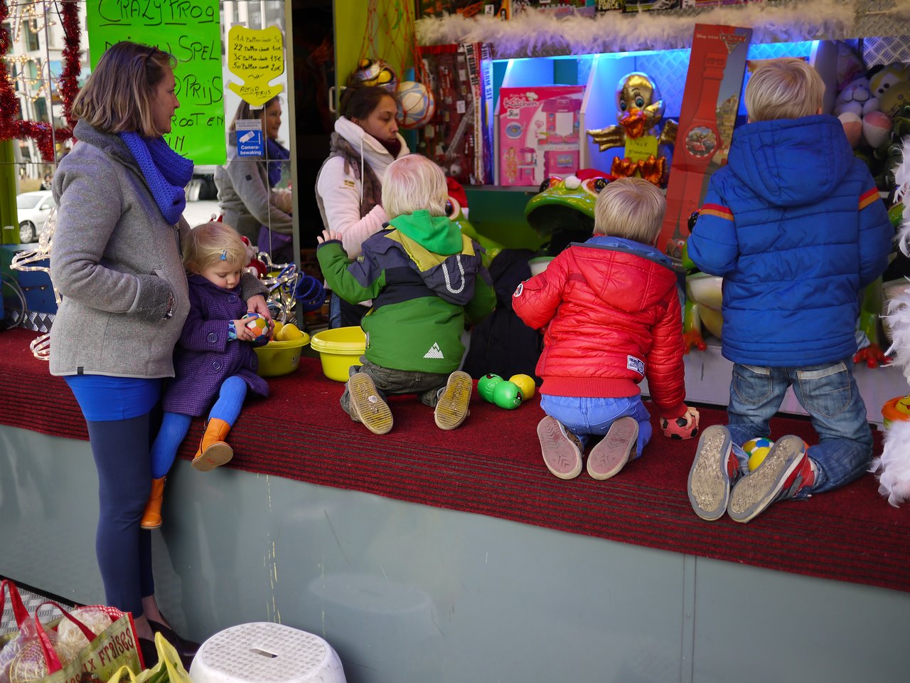 Children in colorful jackets kneel at a toy counter while a woman holds a young child nearby.