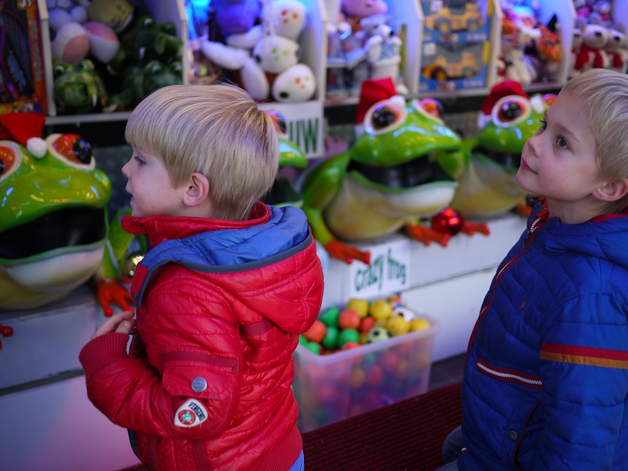 Two young boys in colorful jackets look at large green frog toys in a store.